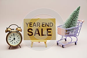 Year end sale text message with wooden easel and alarm clcok, christmas tree on trolley shopping cart on pink background