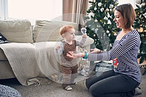 1 year old baby boy standing on the floor near his mother. the first steps concept. christmas decorations on a background