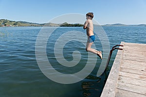 A 11-year-old boy dives into the blue water of a large lake from a wooden pier on a summer sunny day