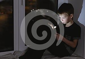 7year old boy looking something on tablet. Sit on windowsill in the dark at night.