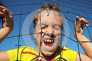 An 8-year-old Caucasian girl stands behind a sports net and screams. Volleyball sports dividing net against the blue sky
