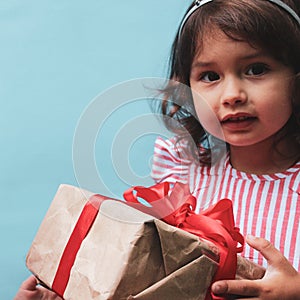 A 3-year-old girl in a red and white dress with a gift in her hands. The gift is packed in kraft paper with a red ribbon
