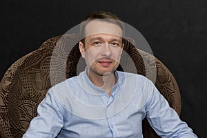 A 30-35-year-old man in a blue shirt sits in an armchair and communicates on a black background. Perhaps he is a lawyer or a lawye