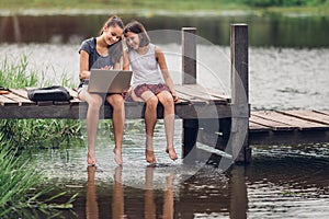 A 13-year-old sister and her 11-year-old sister sit Teach homework with a computer with an internet on a wooden bridge, The river