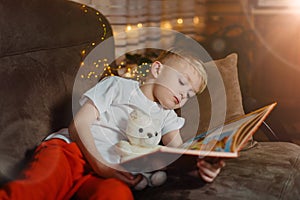 3-4 years old boy is reading a book sitting on a sofa with teddy bear. Home reading of fairy tales. Cute little boy is