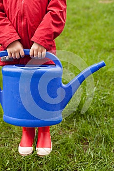 4 years old boy in a red jacket and rubber boots is going to water a tree and from a nice big blue watering can.