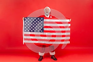 Yelling elderly man with gray beard wearing santa claus costume showing USA flag and screaming, celebrating enjoying Christmas