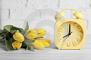 Yellow alarm clock and beautiful tulips on white wooden table against brick wall. Spring time