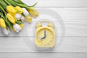 Yellow alarm clock and beautiful tulips on white wooden table, flat lay. Spring time