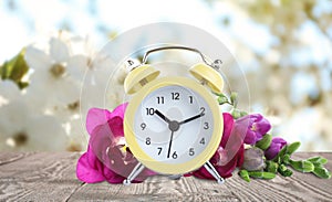 Yellow alarm clock and flowers on table against background. Spring time