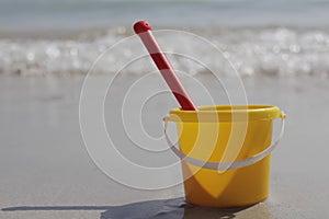 A yellow baby bucket and a red shovel stand on the sand by the sea