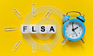 On a yellow background, a blue alarm clock, paper clips and white cubes on which the text is written - FLSA
