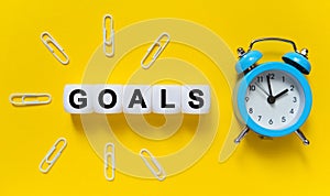 On a yellow background, a blue alarm clock, paper clips and white cubes on which the text is written - GOALS