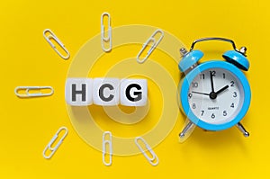 On a yellow background, a blue alarm clock, paper clips and white cubes on which the text is written - HCG