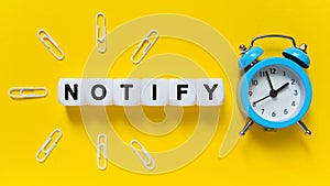On a yellow background, a blue alarm clock, paper clips and white cubes on which the text is written - NOTIFY