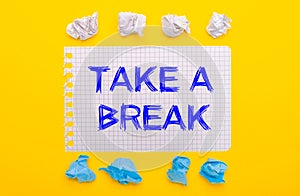 On a yellow background, white and blue crumpled pieces of paper and a notebook with the text TAKE A BREAK