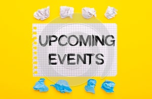 On a yellow background, white and blue crumpled pieces of paper and a notebook with the text UPCOMING EVENTS