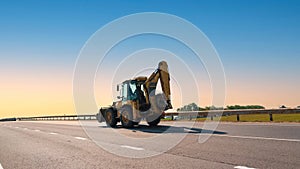 Yellow backhoe loader traveling on highway under clear blue sky signifying ongoing construction work. Open road and photo