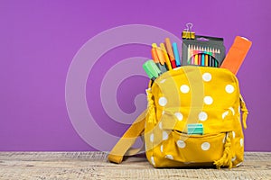 Yellow backpack with white polka dots with different colorful stationery on table. Purple background. Back to school