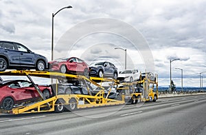 Yellow big rig car hauler semi truck transporting cars on two level hydraulic semi trailer driving on the wide highway road