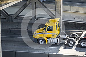 Yellow big rig day cab semi truck with flat bed semi trailer running under the bridge over the divided road