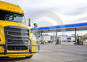 Yellow big rig semi truck with grille guard standing on the truck stop with fuel station on the parking lot