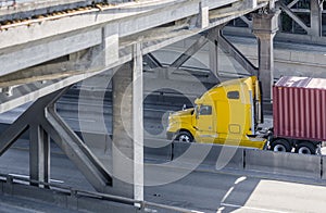 Yellow big rig semi truck transporting container with cargo on flat bed semi trailer running on the highway going under the bridge