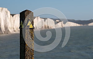 Yellow and black danger sign warning of erosion at the cliff edge. In the background Seven Sisters chalk cliffs, Seaford UK.