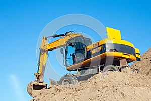 A yellow black excavator standing on a large pile of ground against a blue sky. Digger with a bucket. Road work on a highway