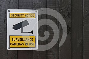 Yellow, black and white Security Notice, Surveillance Cameras In Use warning sign on a painted wooden fence