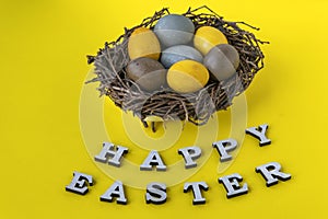 Yellow blue and brown Easter eggs in birds nest on yellow background. Happy easter lettering. Top view