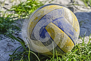 The yellow-blue volleyball ball is old, scratched lying on the sand at the edge of the grass, illuminated by the evening