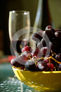 A Yellow bowl of ruby-red grapes, with a wine glass and bottle in the background. Still life with grapes