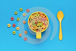 A yellow bowl, a yellow spoon and a breakfast of multicolored corn rings on a blue background. Flat lay