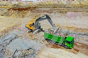 Yellow bulldozer mines clay and loads into green truck.