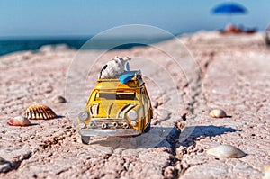 Yellow car toy with surfboard and seashell driving on a stone road along the sea coast