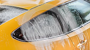 Yellow car washed in self serve carwash, shampoo spraying from hose to roof, streams of foam flowing down the window