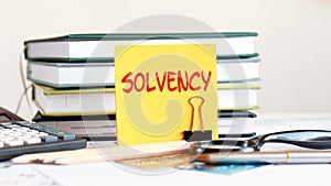 A yellow card with the text solvency stands on a clip for papers on the desk against the background of books, selective focus