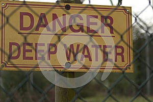 Yellow caution sign with the message Danger, Deep Water written in red behind the metal fence