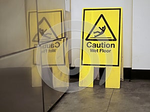 Yellow Caution wet floor sign by a wall reflects in a stainless fridge in a canteen of supermarket. Accident prevention and