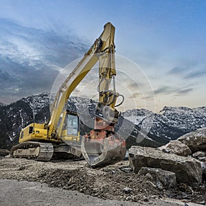 yellow chain excavator with grapple of natural stone in front of a mountain backdrop
