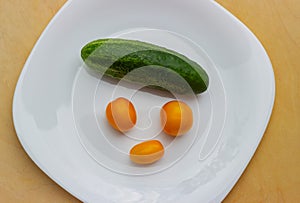 Yellow cherry tomatoes, ripe cucumber on a white glossy plate in the form of a sad facial expression