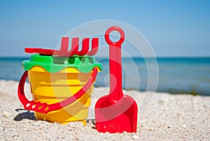 Yellow baby bucket with red handle, plastic red spatula and rake, and plastic green sieve, left on a background of blue sea sand s