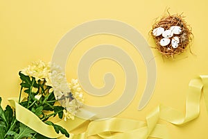 Yellow chrysanthemums flowers bouquet with beautiful wide ribbon and nest with Easter eggs on yellow background. Greeting card