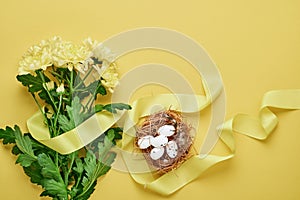 Yellow chrysanthemums flowers bouquet with beautiful wide ribbon and nest with Easter eggs on yellow background. Greeting card