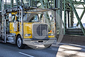 Yellow classic bonnet big rig semi truck tractor with empty two level hydraulic semi trailer driving on the truss arched bridge