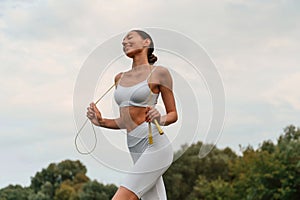 Yellow colored jumping rope. Young caucasian woman with slim body shape is in the fitness clothes outdoors