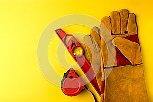 Yellow construction protective gloves, red building level and tape measure on yellow background.