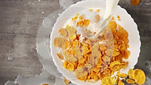 Yellow cornflakes and milk are pouring and mixing in slowmo into a white plate