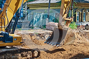 Yellow crawler excavator at a road reconstruction site in a village with old log houses. Summer sunny day. No people